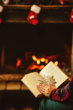 Woman reading a book by the fireplace. Young woman reading a book by the warm fireplace decorated for Christmas. Relaxed holiday evening concept.の写真素材