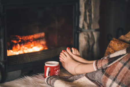 Woman relaxes by warm fire with a cup of hot drink and warming up her feet.の写真素材
