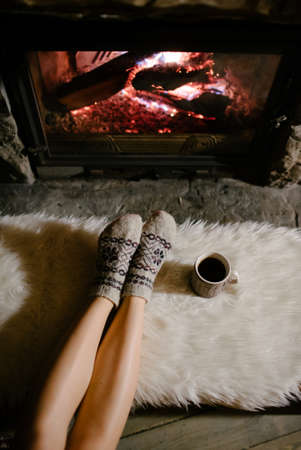 Woman relaxes by warm fire with a cup of hot drink and warming up her feet in woolen socks.