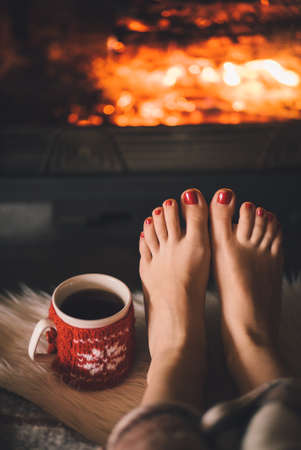 Woman relaxes by warm fire with a cup of hot drink and warming up her feet.の写真素材