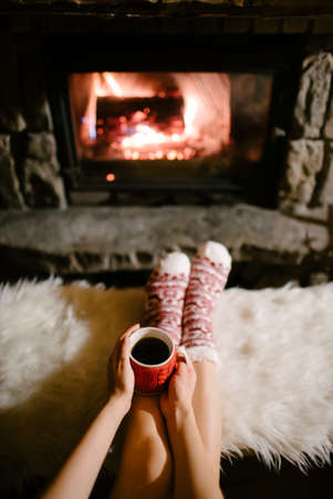 Woman relaxes by warm fire with a cup of hot drink and warming up her feet in woolen socks.の写真素材