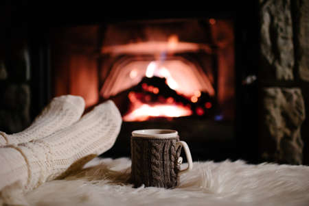Woman relaxes by warm fire with a cup of hot drink and warming up her feet in woolen socks.の写真素材