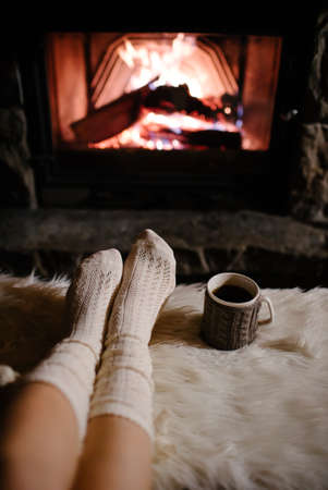 Feet in woollen socks by the Christmas fireplace. Woman relaxes by warm fire with a cup of hot drink...