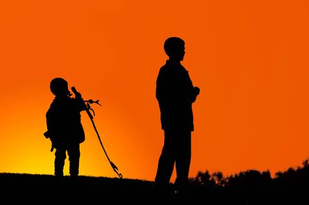 Two boys standing on a hill dark silhouette over colorful digitally adjusted orange evening skyの写真素材