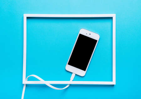White cell phone on a blue background in a frame. Concept of black screen, black mirror. a picture of the modern world, where people are ruled by technologyの写真素材