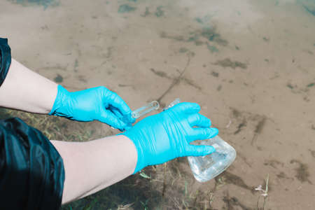 Fence, check the water sample for infections. Gloved hand with a flask and a test tube on a background of lake, river, sea. Pollution of ecology, environmentの写真素材
