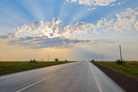 An asphalt road in the middle of a field with the rays of the sun through the cloudsの写真素材