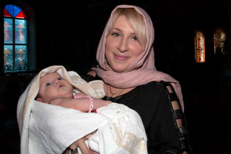 A woman with a baby in her arms in a darkened part of an Orthodox church. The sacrament of baptism...