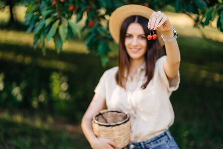 Portrait of a young cheerful woman in a straw hat with a basket, harvests sweet cherries in the...
