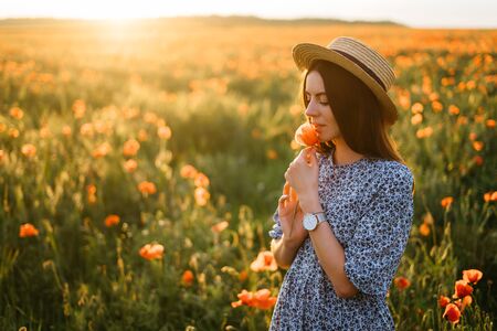 Amazing young woman in hat and blue dress posing in a poppies field. Beautiful happy tender young woman enjoying the smell of summer wildflowersの写真素材