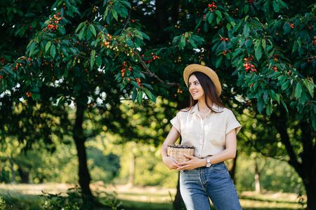Portrait of a young cheerful woman in a straw hat with a basket, harvests sweet cherries in the...