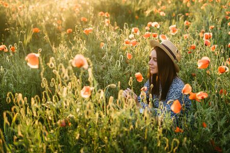 Amazing young woman in hat and blue dress posing in a poppies field. Beautiful happy tender young woman enjoying the smell of summer wildflowersの写真素材