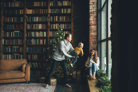 Happy family in cozy living room with decorated Christmas tree. Young father is throwing his little son up, a beautiful mother is laughingの写真素材