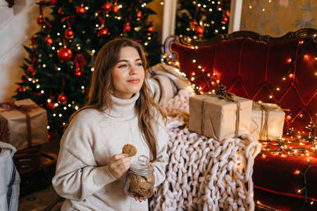 Young woman smiling, relaxing at home, enjoying Christmas. Beautiful girl eats cookies sitting near the Christmas treeの写真素材