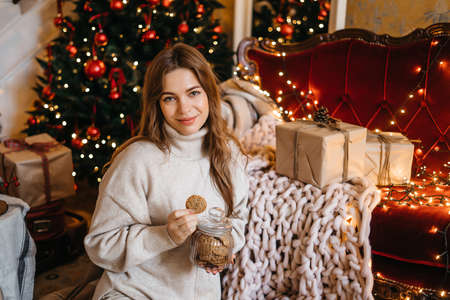 Young woman smiling, relaxing at home, enjoying Christmas. Beautiful girl eats cookies sitting near the Christmas treeの写真素材