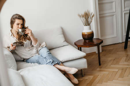 A young blonde woman in casual comfortable clothes sits on a white sofa at home and drinks coffee. scandinavian interior, minimalismの写真素材