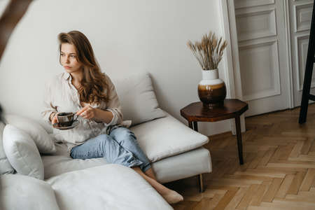 A young blonde woman in casual comfortable clothes sits on a white sofa at home and drinks coffee. scandinavian interior, minimalismの写真素材