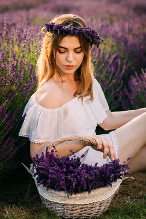 A young and happy blonde woman in a delicate dress is enjoying the spring in a lavender field at sunset, holding a basket full of flowers in her hands. High quality photoの写真素材