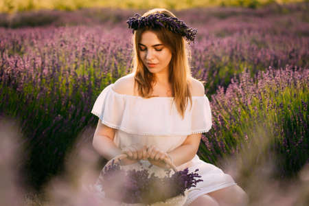 A young and happy blonde woman in a delicate dress is enjoying the spring in a lavender field at sunset, holding a basket full of flowers in her hands. High quality photoの写真素材
