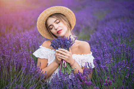 Young blond woman in lavender field. Happy carefree female in a white dress and straw hat enjoying sunset. Outdoors portrait. High quality photoの写真素材