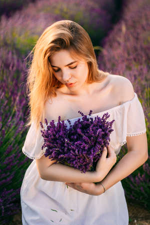 Young and happy blond woman and white dress enjoying spring in a lavender field at sunset holding a bouquet of flowers in her hands. High quality photoの写真素材