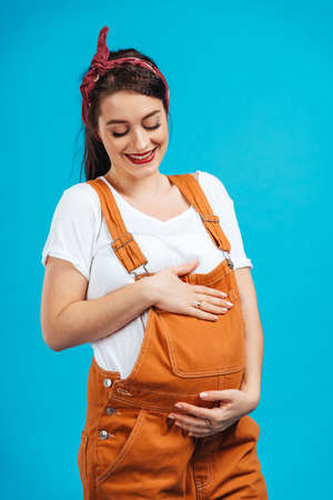 Cheerful pregnant brunette woman touching pregnant belly smiling, dressed in pin-up style, isolated...