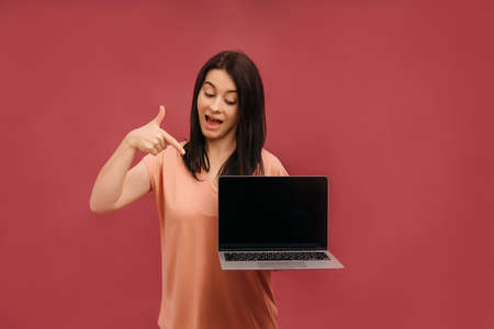 Portrait of a cheerful young casual girl standing isolated over pink background, pointing at laptop computer. High quality photoの写真素材