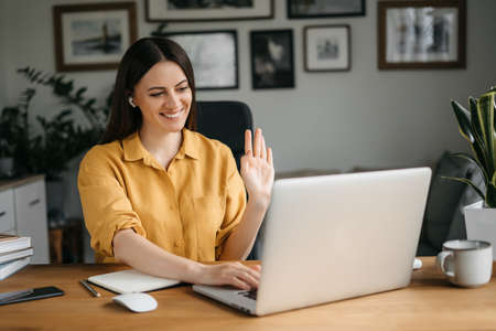 Young pretty woman talking on video call and waving hand while sitting at table in the homeoffice room. High quality photoの写真素材