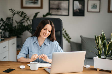 Young pretty woman talking on video call and waving hand while sitting at table in the homeoffice room. High quality photoの写真素材