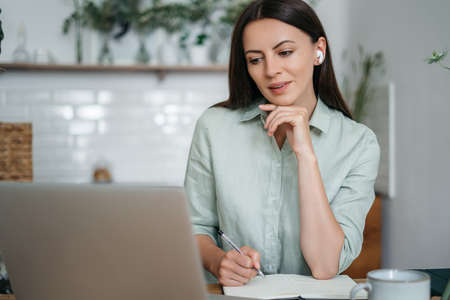 Millennial girl sit at desk in living room study on laptop making notes, concentrated young woman...