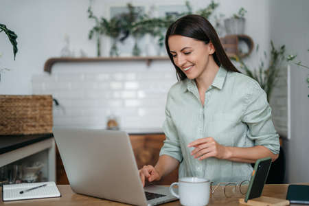 Young pretty woman talking on video call and waving hand while sitting at table inthe kitchen. High quality photoの写真素材