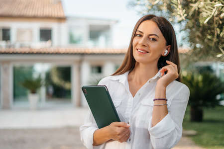 Portrait of a happy brunette woman smiling holding a tablet in the hands of a summer garden white shirt. The realtor presents and sells the house. High quality photoの写真素材