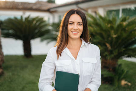 Portrait of a happy brunette woman smiling holding a tablet in the hands of a summer garden white shirt. The realtor presents and sells the house. High quality photoの写真素材