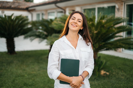 Portrait of a happy brunette woman smiling holding a tablet in the hands of a summer garden white shirt. The realtor presents and sells the house. High quality photoの写真素材