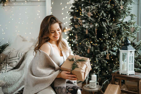 Smiling sincere beautiful young woman in light clothes holds out a wrapped gift box to the camera, prepares a Christmas present, celebrates the winter holidays, sitting on a sofa near a decorated treeの写真素材