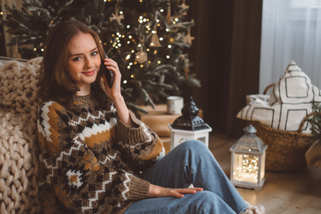 A charming young red-haired woman on the background of a Christmas tree in a cozy living room holds a mobile phone and waves her hand talks with her friend or family via video call. High quality photoの写真素材