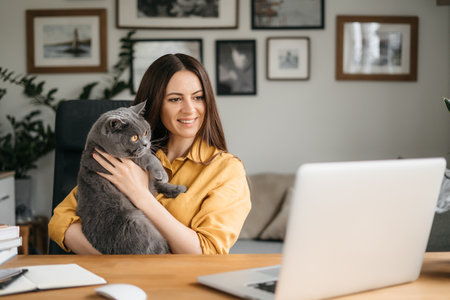 Beautiful young woman smiling on video call via laptop computer, sitting at table at home talking online with family and friends, holding british cat in her arms. High quality photoの写真素材