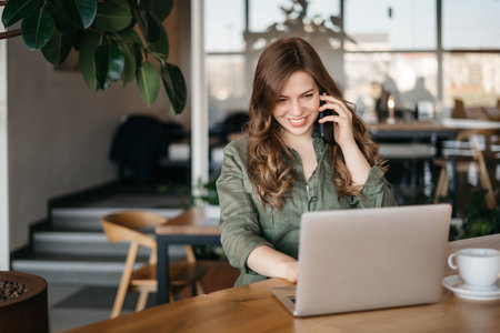 Woman books tickets for concert. Portrait of good-looking caucasian girl sitting in cafe, drinking coffee, talking on smartphone, looking at laptop screen with broad smile, checking profile of friend.の写真素材