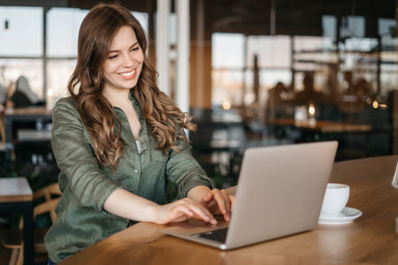 Happy Latin Project Manager with Dark Wavy Hair Working on Laptop in Busy Creative Office. Beautiful Diverse Female Ad Manager Comparing Reports and Graphs on Paper and on Her Computerの写真素材