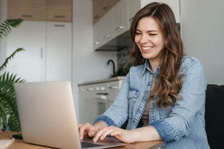 Smiling young woman working on laptop computer while sitting on a kitchen, holding mobile phone. High quality photoの写真素材