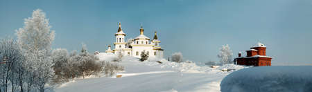White orthodox church amidst deep snow and bare trees on the background of clear blue sky panoramaの写真素材