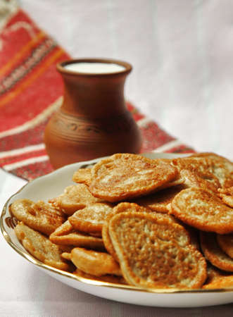 Heap of potato flapjacks, clay jug with milk and bowl with sour cream on the tablecloth with national pattern close upの写真素材