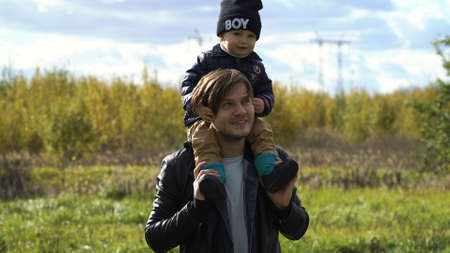 Happy father and little son walking in autumn park and having fun.Son sits on his fathers neck.Happy young family spending time together outside in green nature.Happy family enjoying life together at meadow outdoor.の写真素材