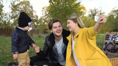Smiling family taking selfie sitting on the grass in a city park. Young family with son at a picnic...