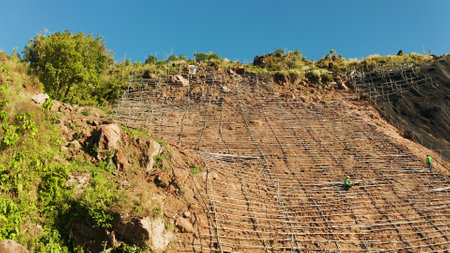 Workers strengthen the slope of the mountain with metal mesh preventing rockfall and landslide on the road, above view. workers constructing anti-landslide concrete wall prevent protect against rock slides. safety conceptの写真素材