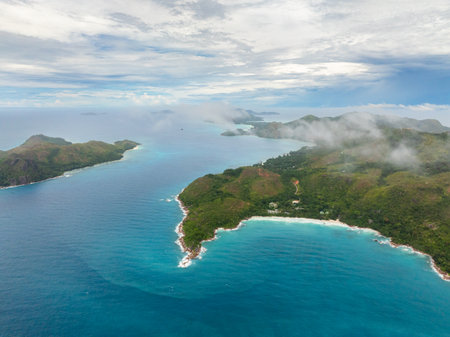 Aerial view of lush green hills shrouded in mist, with turquoise waters and a rocky coastline stretching into the horizon. Praslin, Seychelles.の写真素材