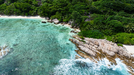 Granite formations meet clear turquoise waters along a white sandy beach surrounded by greenery....
