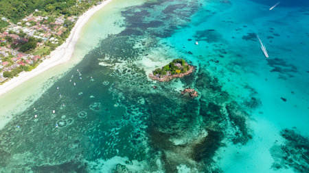 A small, lush island sits in shallow turquoise waters, surrounded by coral reefs and close to a...