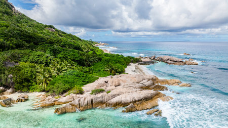 Waves break over granite rocks along a coastline fringed by lush greenery and clear waters. Seychelles, La Digue.の写真素材