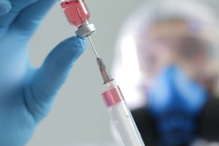 A doctor in a protective suit blue gloves with a respirator draws a vaccine from a vial into a syringeの写真素材